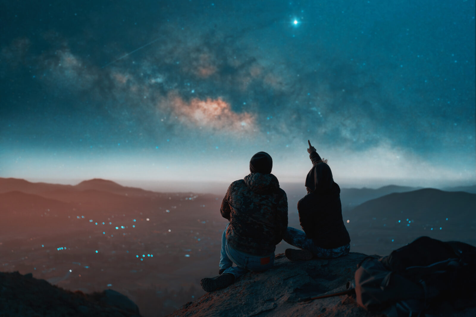 silhouette of a couple sitting on top of a hill looking at the stars over the city, back view