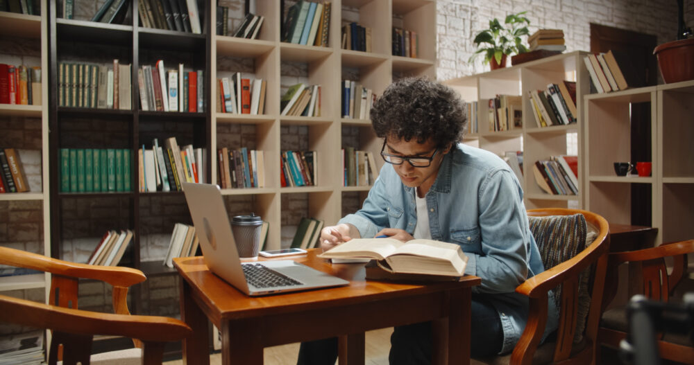 Asian student doing research in library using laptop in books, making a project and preparing for exam. Nerdy guy studying his lessons - education, self-study concept close up