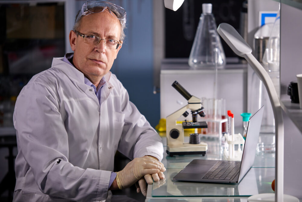 senior scientist man in white dress coat sitting in laboratory at work place, successful serious face and feeling expression. caucasian microbiologist make research