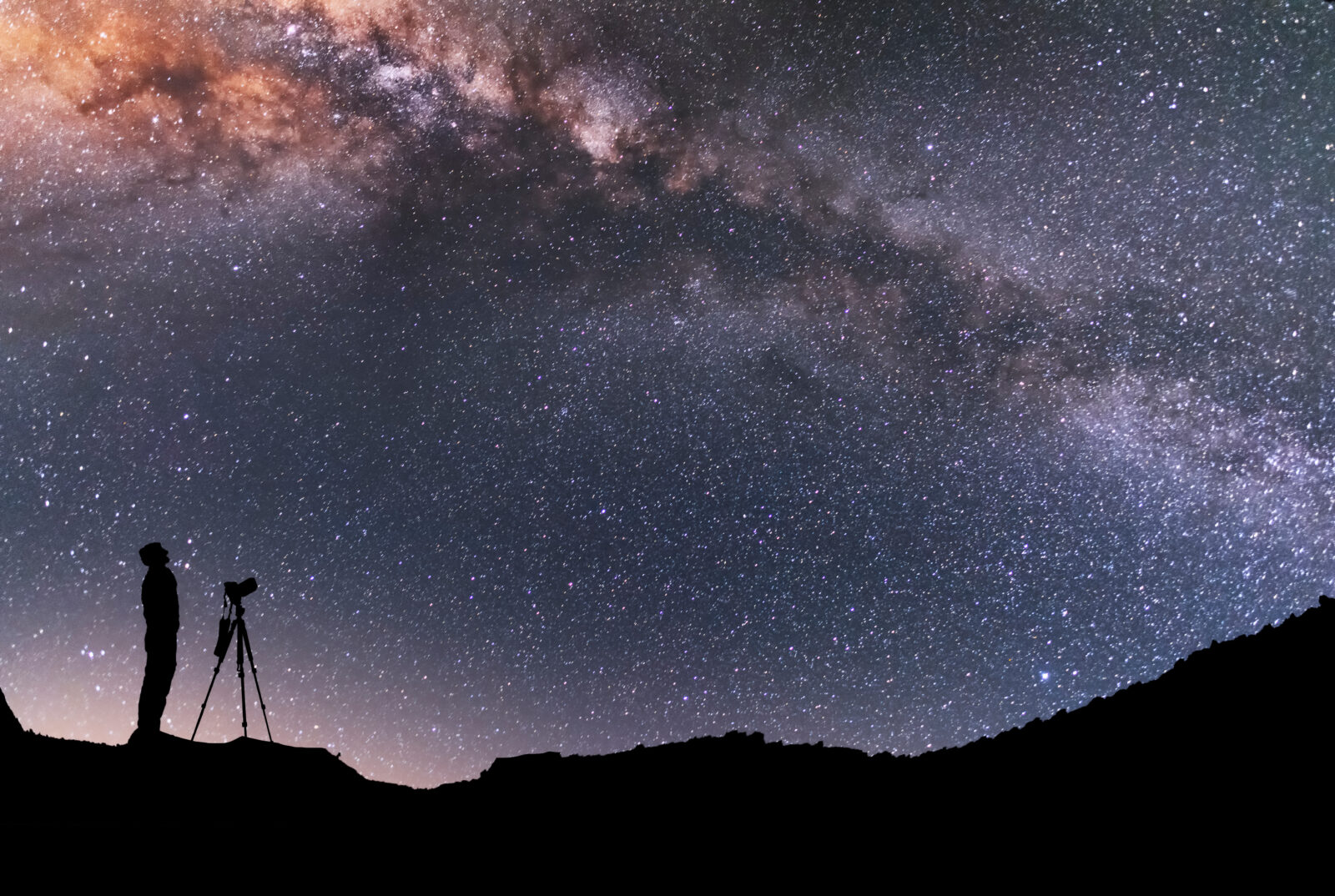 The man silhouette with camera and tripod, stands on the hill and looking at the bright milky way galaxy. Beautiful night landscape.