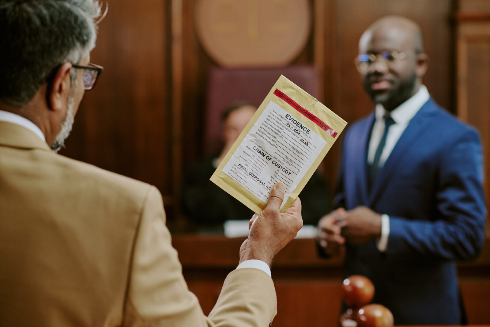 Middle aged Caucasian man holding evidence bag while standing in courtroom, Black man in suit standing in background, both participating in legal proceedings, judge visible behind